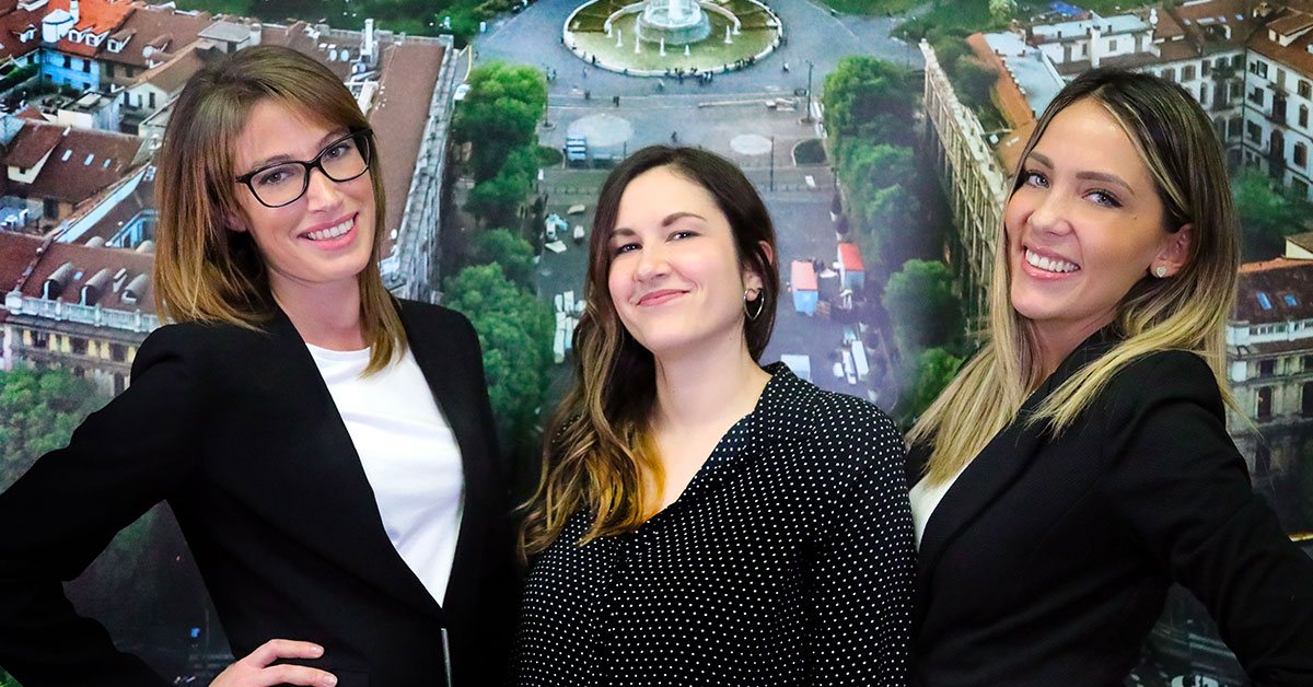 Tre persone dello staff sorridono davanti a una grande foto aerea di Milano